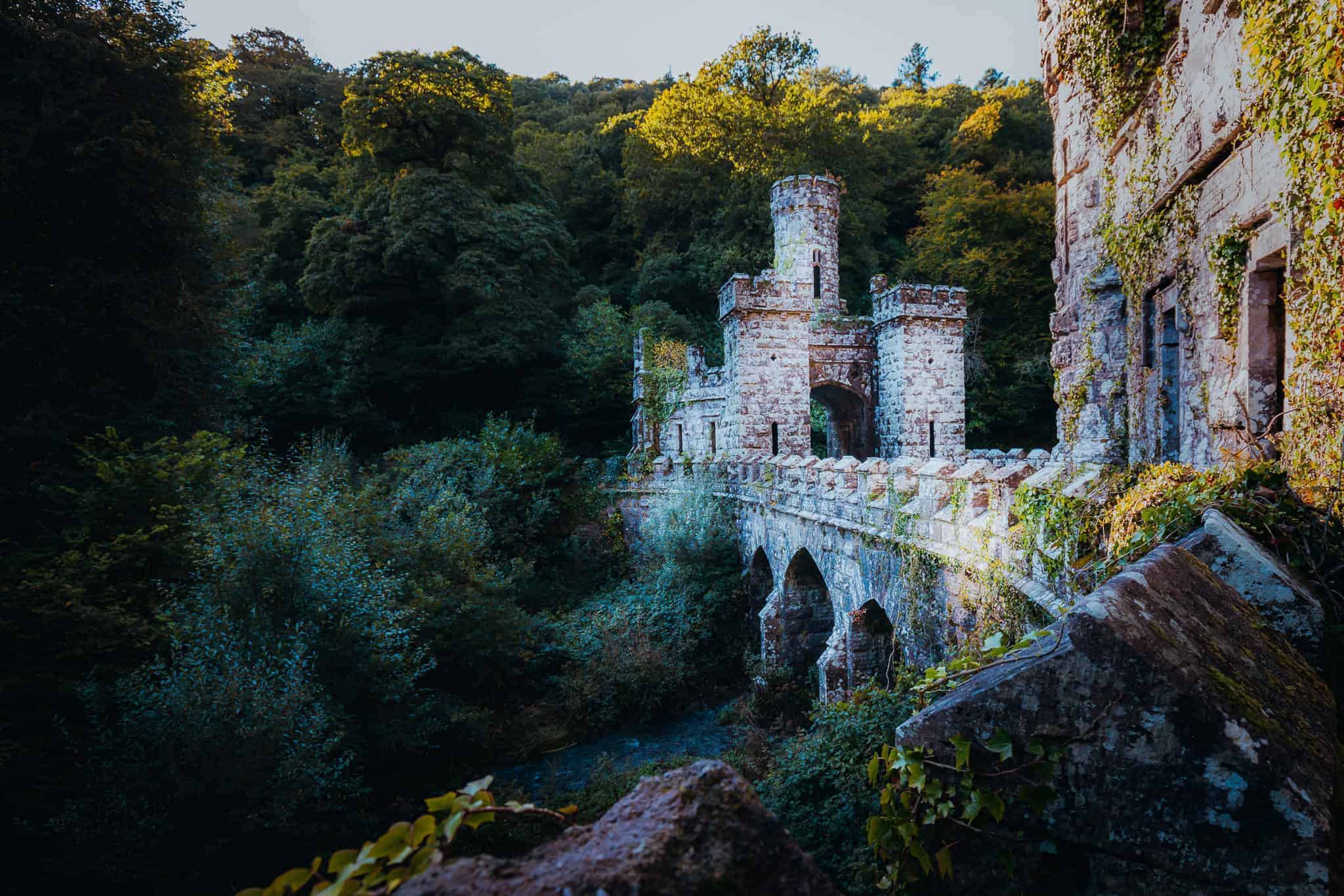 A picturesque view of Ballysaggartmore Towers, showcasing its stone bridge with arched supports leading to a medieval-style tower nestled among dense, green forest. Ivy and foliage partially cover the weathered stone structure, with soft sunlight filtering through the trees, creating a mystical atmosphere.