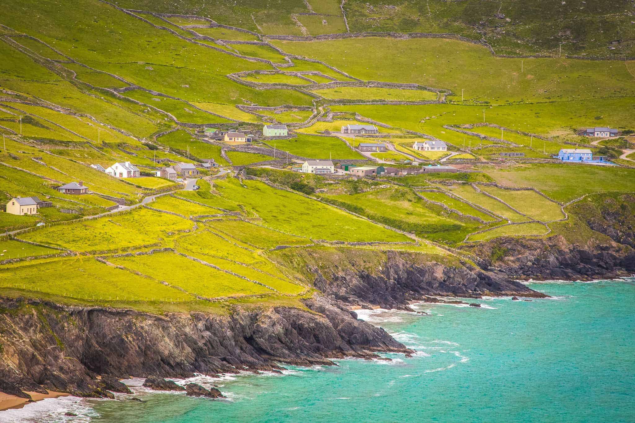 A scenic coastal view of a rural Irish village with scattered houses, winding stone walls, and green pastures overlooking the turquoise Atlantic Ocean.