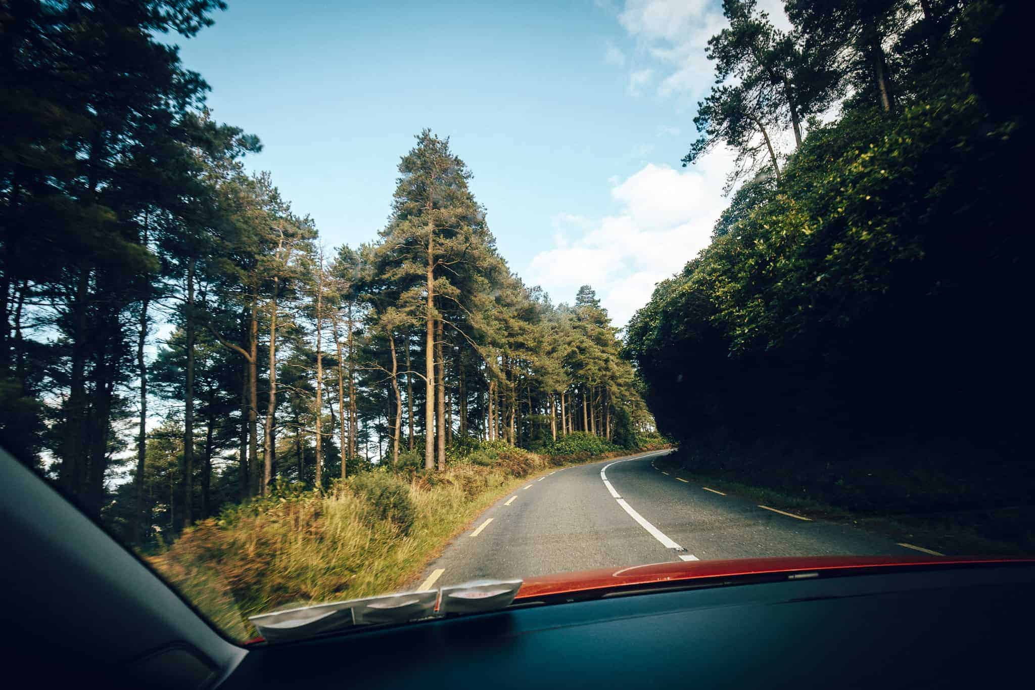 View from inside a car driving along the scenic Vee Pass in Ireland. The narrow winding road is lined with tall pine trees on the left and dense greenery on the right under a bright blue sky with scattered clouds. The car’s red hood and dashboard are partially visible in the foreground.