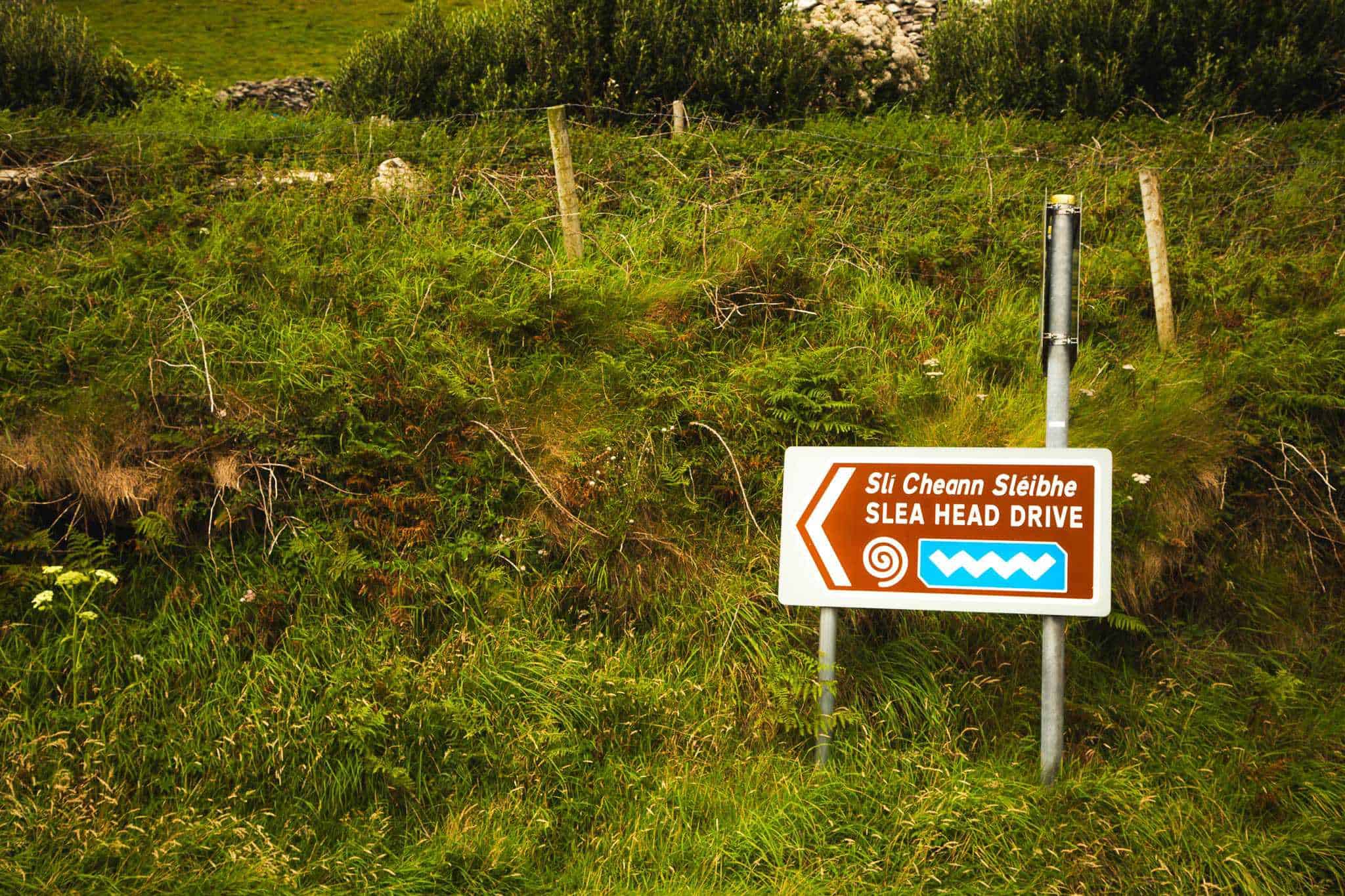 A brown and white directional road sign reading "Sli Cheann Sléibhe Slea Head Drive" with a spiral and zigzag symbol, set against a lush green roadside.