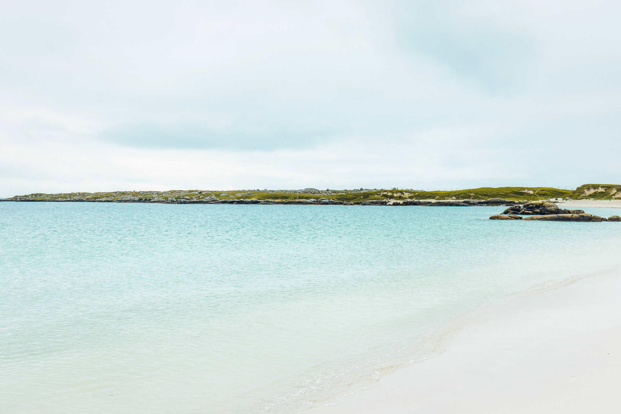 Calm turquoise waters of Dog’s Bay gently meeting the soft white sand, with low grassy hills across the horizon.