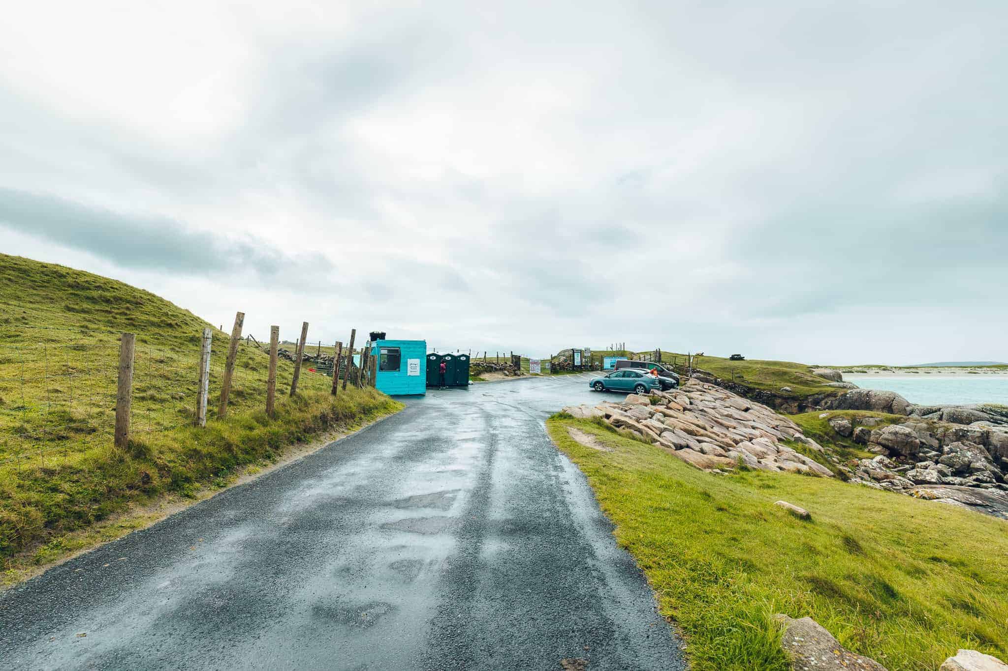 Small coastal parking area with cars, portable toilets, and a turquoise shed beside grassy hills and rocky shoreline.