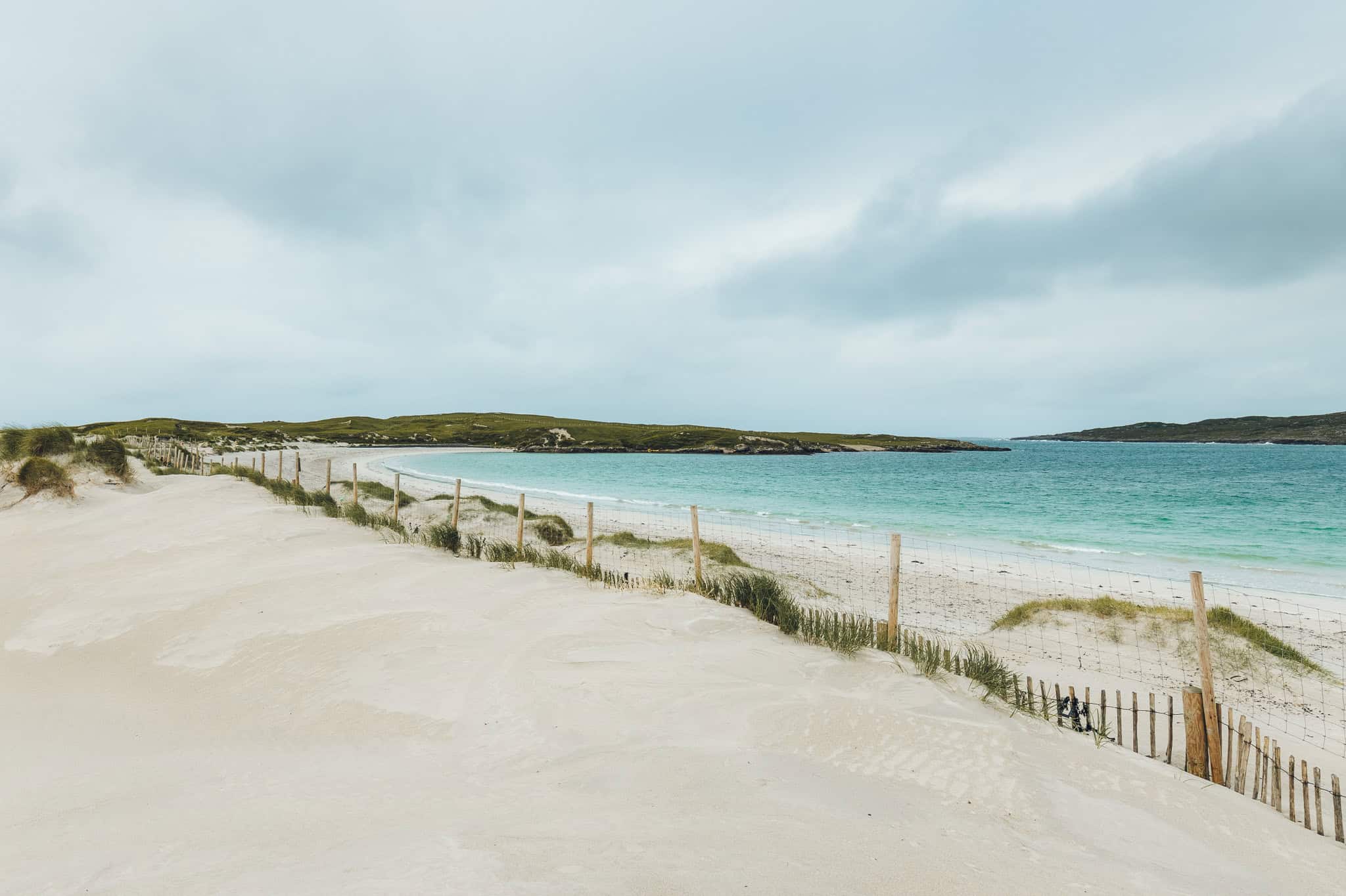 Soft white sand dunes fenced off above Dog’s Bay, with turquoise waters and grassy hills in the distance.