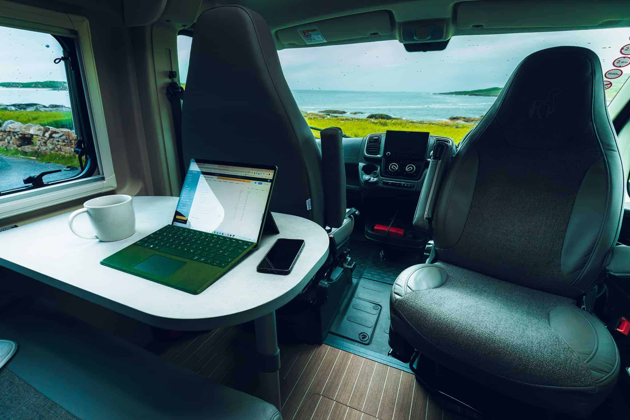 Inside a campervan showing a table setup with a laptop, coffee mug, and ocean view out the window.