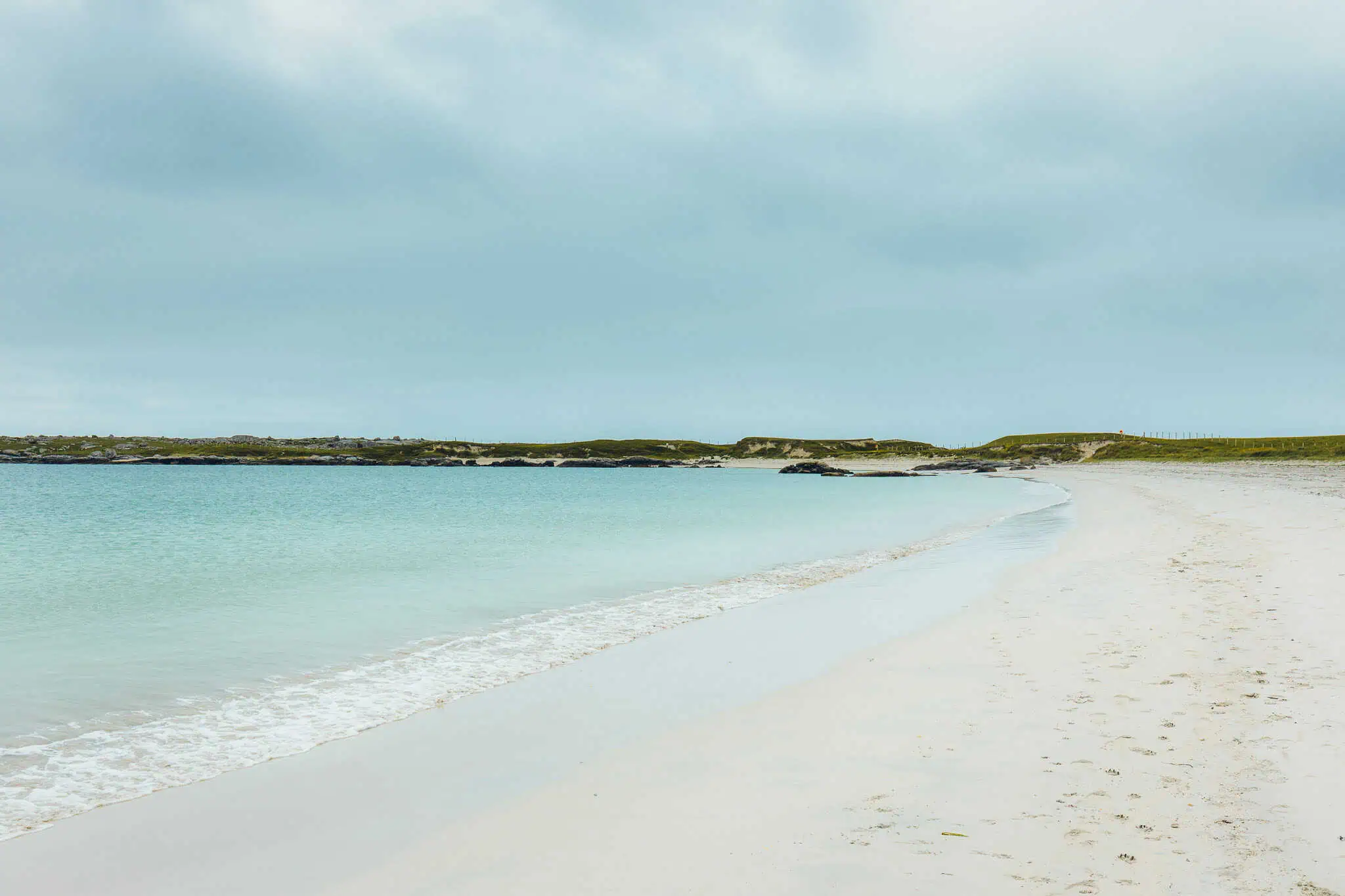 Peaceful stretch of soft white sand curving around calm turquoise water under an overcast sky.