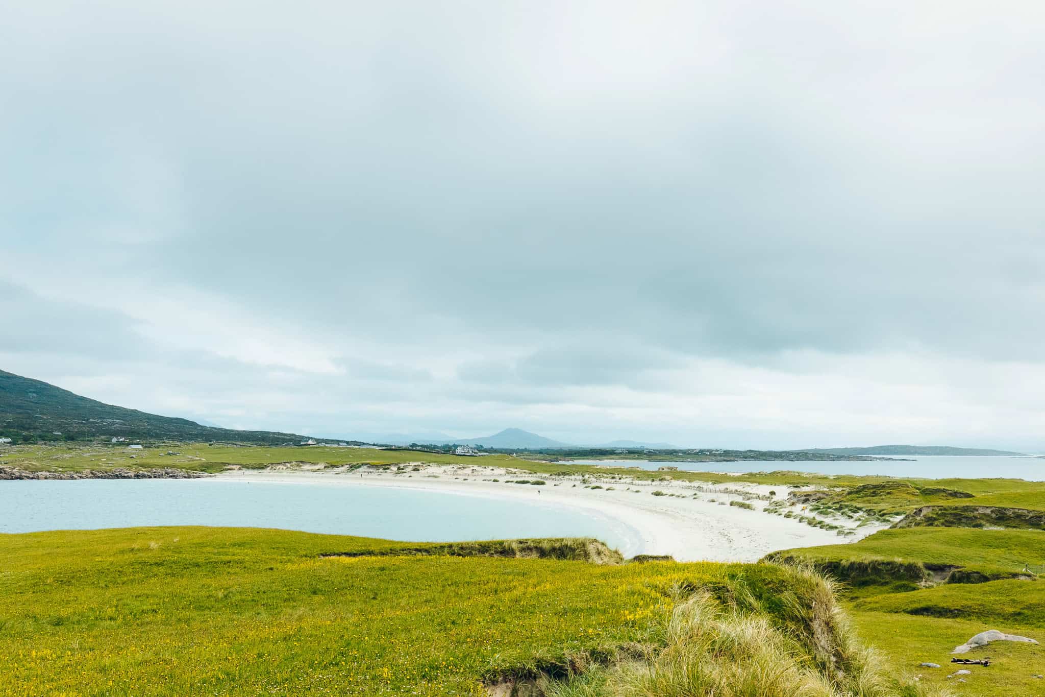 Wide view of Dog’s Bay and surrounding green hills, with a distant mountain rising under moody clouds.