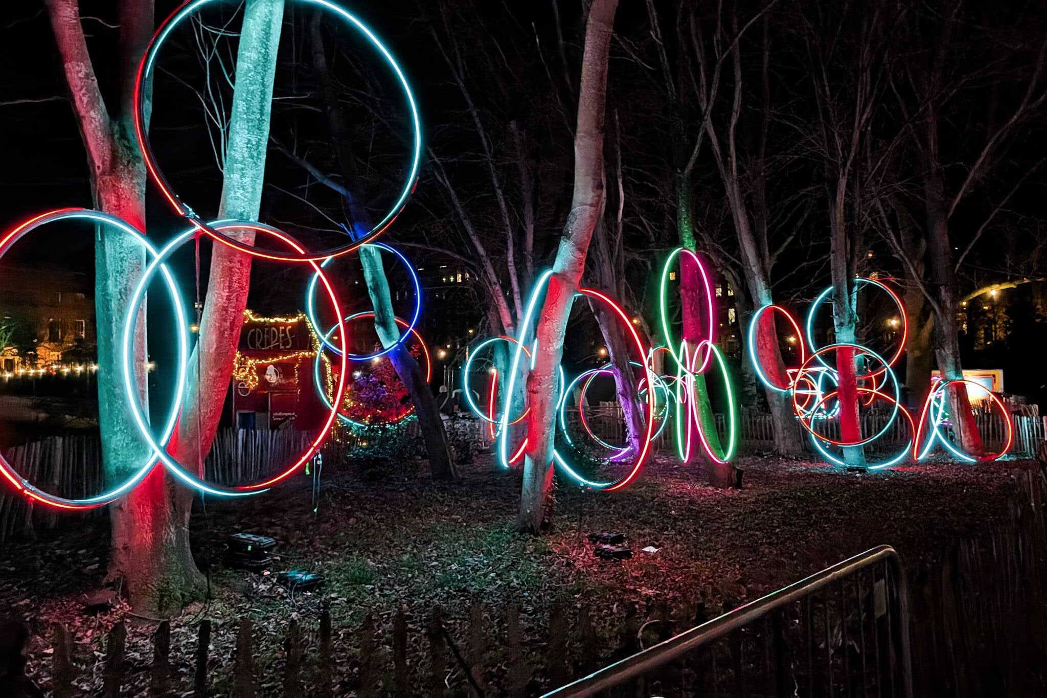 Colorful neon hoops in red, blue, green, and purple glow among the trees at the Merrion Square Winter Lights event in Dublin. A small crepe stand lit with festive garlands is visible in the background, adding to the whimsical nighttime atmosphere.