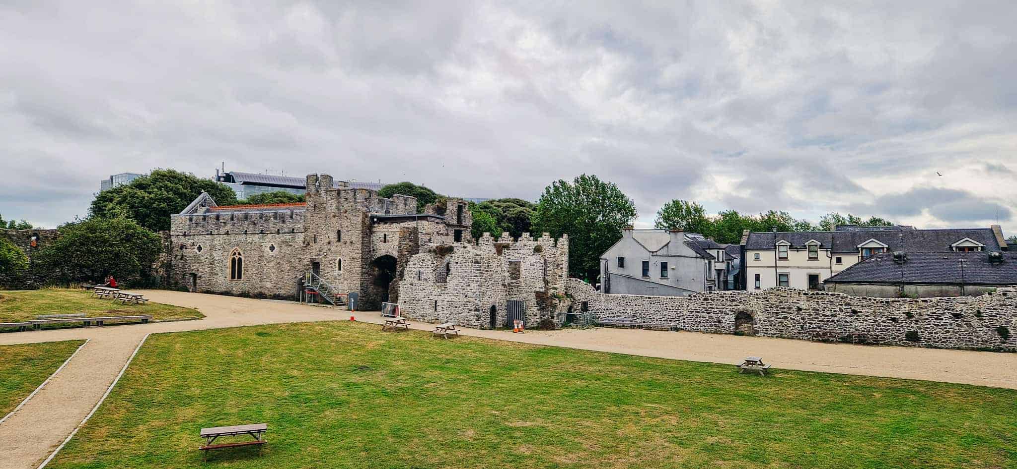 A wide view of Swords Castle's grassy courtyard surrounded by stone walls, with picnic benches scattered around and modern buildings visible beyond the castle's perimeter.