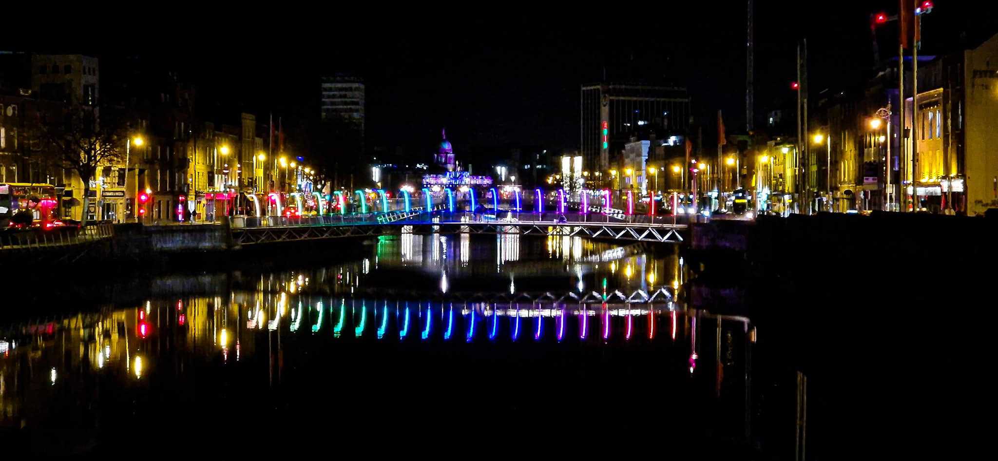 A vibrant night view of Dublin’s Millennium Bridge lit with colorful lights in shades of blue, green, red, and yellow, reflecting beautifully on the calm waters of the River Liffey.