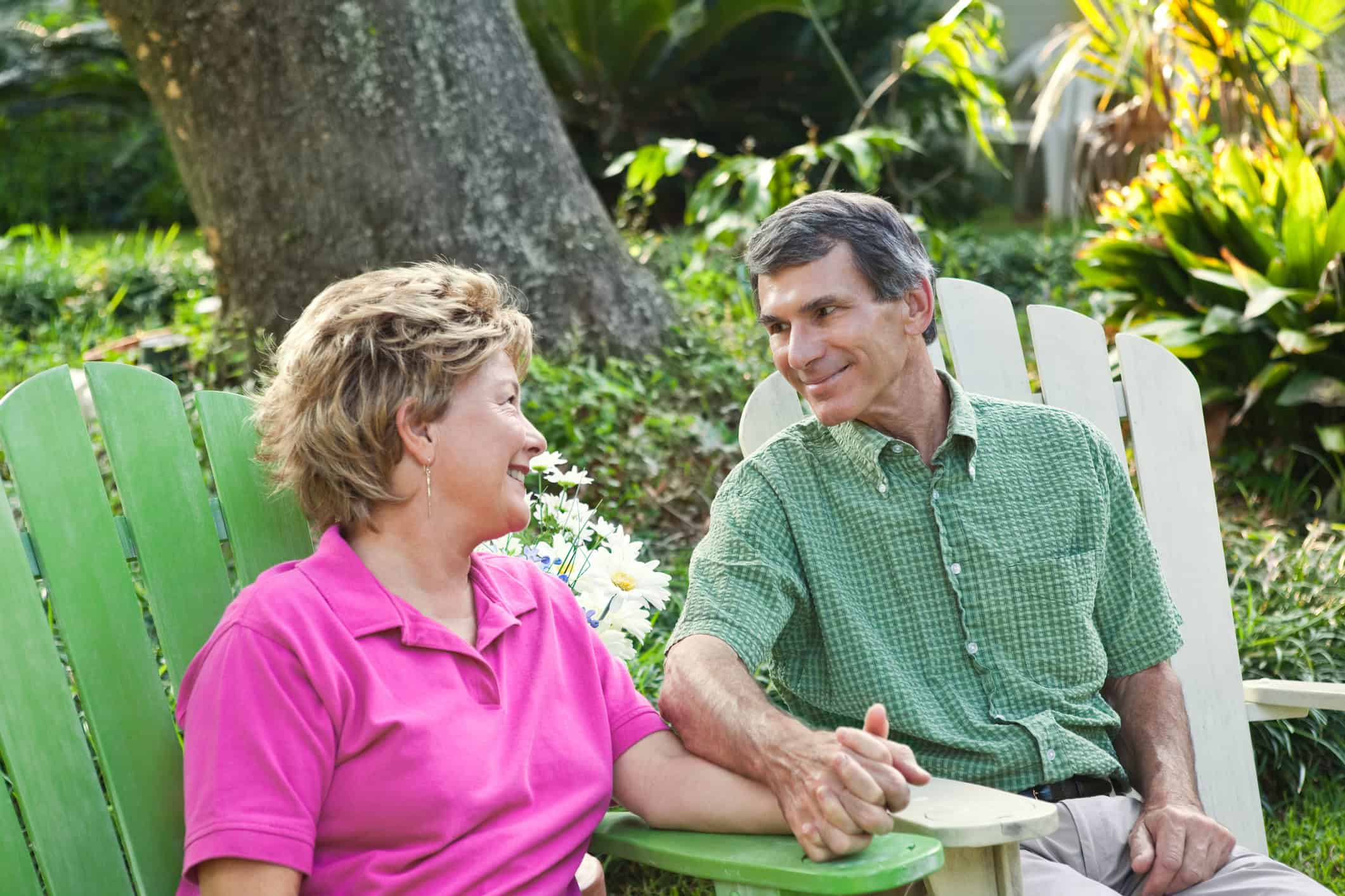 A mature couple sitting in Adirondack chairs holding hands and gazing at each other with a lasting love