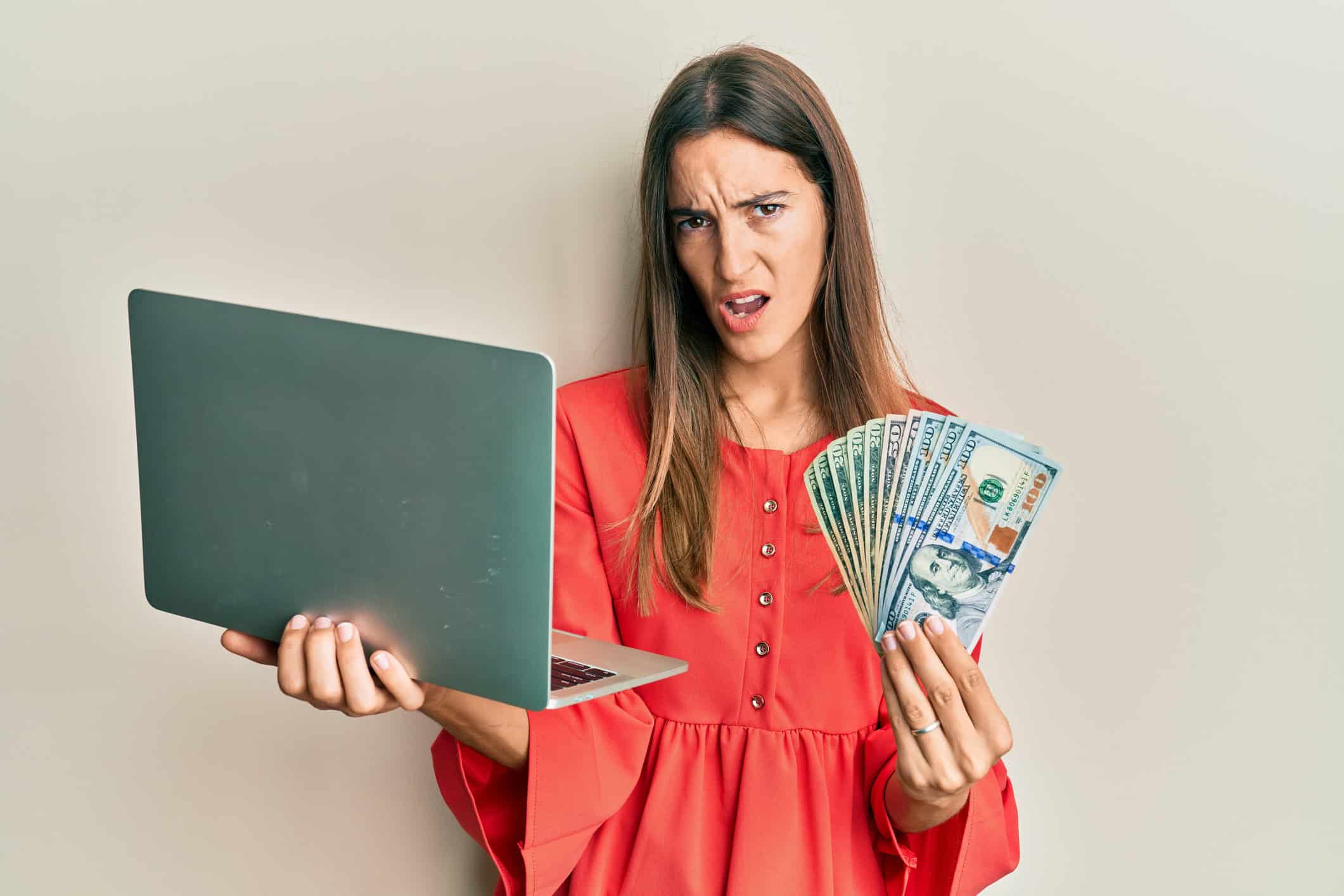 Woman holding a laptop and $100 bills looking shocked.
