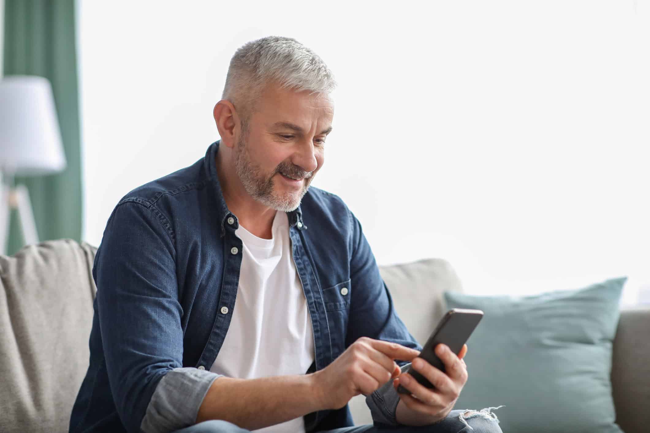 A handsome, happy man sitting on his couch and reading an article on his smart phone about online dating help for men.