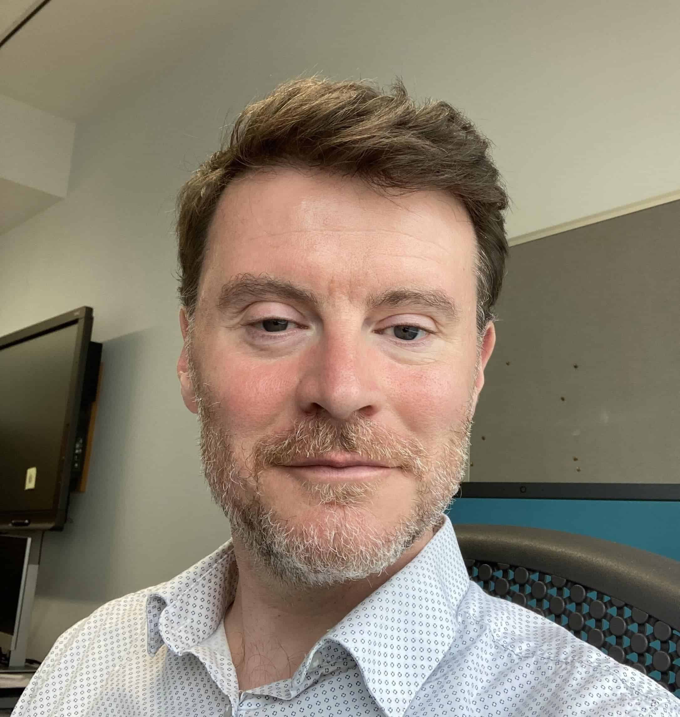 Man with brown hair and beard in office environment, casual business attire, close-up portrait for professional profile.