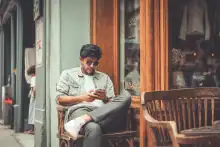 Um homem de óculos escuros e jaqueta jeans senta em um banco de madeira, usando o celular na calçada de um café com vitrine de madeira.
