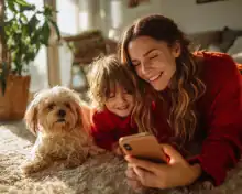 Uma mulher e uma criança sorridentes, vestindo vermelho, deitadas no tapete com um cachorro fofo, olhando para um celular sob a luz do sol.
