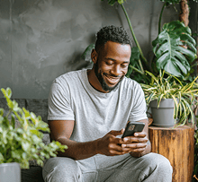 Um homem negro sorridente, vestindo cinza, senta-se em um sofá e olha para seu smartphone, cercado por várias plantas verdes em um ambiente interno aconchegante.