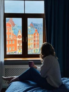 Woman sitting on a hotel bed holding a mug while looking through a window at the colorful historic buildings of Riga Old Town. The view highlights the atmosphere travelers can enjoy when choosing where to stay in Riga.