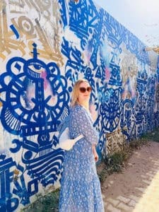 Woman in a light blue patterned dress and sunglasses standing beside a large blue and white mural under bright sunshine. Loose fitting clothing, sun protection, and lightweight layers demonstrate smart ways to stay cool and comfortable when travelling in very hot weather.