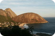 Vista panorâmica de uma enseada com uma praia de areia, casas, montanhas rochosas e o oceano azul com ondas sob um céu claro.