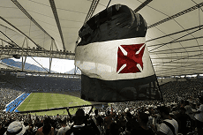 Torcedores de futebol levantam uma gigantesca bandeira preta e branca com uma cruz de Malta vermelha em um estádio moderno e lotado durante um jogo.