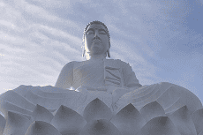 Uma grande estátua branca de Buda sentado em posição de lótus sobre uma base de flor de lótus, contra um céu azul com nuvens esparsas.
