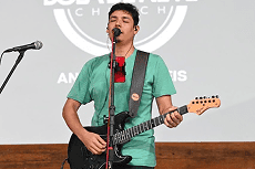 Um jovem de camiseta verde canta com olhos fechados e toca guitarra elétrica preta em um palco, com fundo cinza e logotipo de igreja.