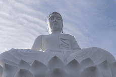 Uma grande estátua branca de Buda sentado em posição de lótus sobre uma base de flor de lótus, contra um céu azul com nuvens esparsas.
