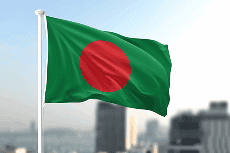 A bandeira de Bangladesh, verde com um círculo vermelho, tremula num mastro branco sob um céu azul claro com um horizonte urbano desfocado ao fundo.