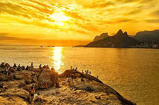 Pessoas se reúnem em rochas para admirar um espetacular pôr do sol dourado sobre o oceano, com montanhas e uma cidade ao fundo.