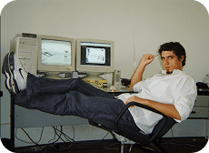 Jovem de camisa branca e jeans relaxa com os pés na mesa de escritório, olhando para a câmera entre dois monitores e um computador antigo.