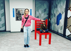 Uma mulher de rosa parece gigante ao lado de um homem pequeno sentado em uma cadeira vermelha enorme, criando uma ilusão de ótica em uma sala geométrica.
