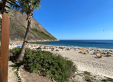 Uma praia ensolarada e movimentada com areia clara, mar azul e muitas pessoas relaxando, ladeada por uma grande montanha rochosa à esquerda.