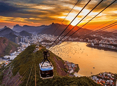 Um bondinho azul desce uma montanha verde, com vista panorâmica da cidade do Rio de Janeiro, uma baía com barcos e um pôr do sol vibrante.