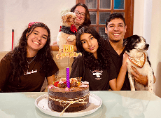 Família sorridente de cinco pessoas e dois cachorros celebra um aniversário com bolo de chocolate e topo de bolo "Happy Birthday" em casa.