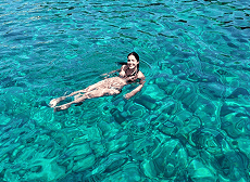 Uma mulher sorridente flutua de costas em águas oceânicas incrivelmente claras e turquesas, com o fundo rochoso visível através da superfície.