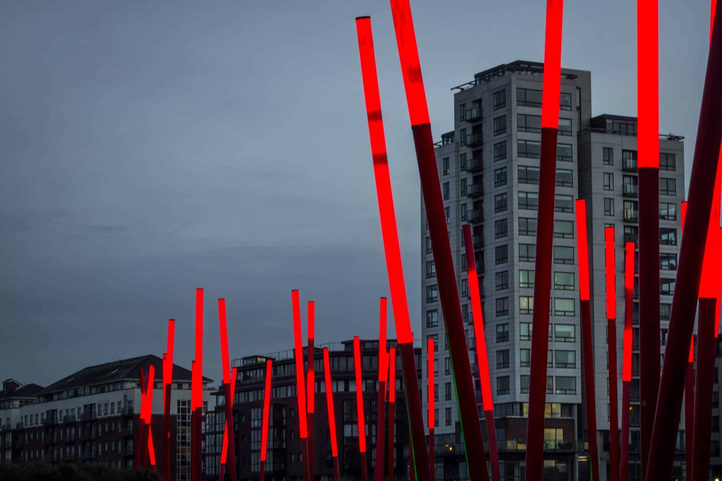 The Grand Canal Dock area in Dublin during twilight, with modern apartment buildings and the iconic red illuminated poles of the Docklands reflecting in the water.