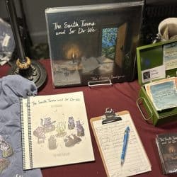 The Earth Turns and So Do We album cover display at a music booth with vinyl records, a spiral notebook, a clipboard with notes, and other music merchandise.
