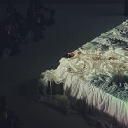 A person lying on a large, flowing white fabric installation at a fashion or art show, surrounded by seated audience members in a darkened venue.
