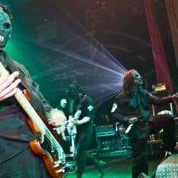 Dark masked guitarist performing live at a concert with dramatic lighting and stage effects, capturing the energy of a heavy metal or rock show.