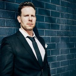 Professional businessman in a tailored suit standing against a modern brick wall, representing insights on the music industry, digital marketing, and entrepreneurship themes on Hypebot.
