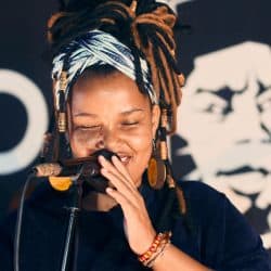 A vibrant female singer with dreadlocks and colorful headwrap performing passionately into a microphone at a music event.