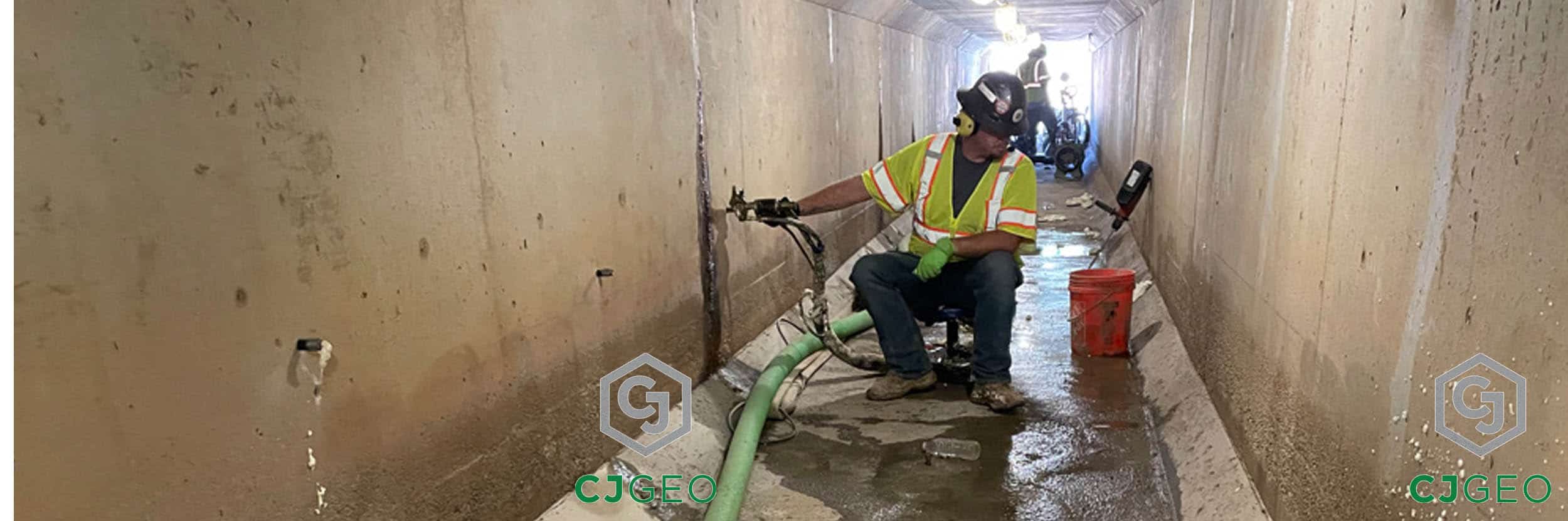 A CJGeo technician in a hard hat and safety vest works from inside a long, concrete box culvert, injecting polyurethane grout into the culvert wall to seal joints and prevent leaks.
