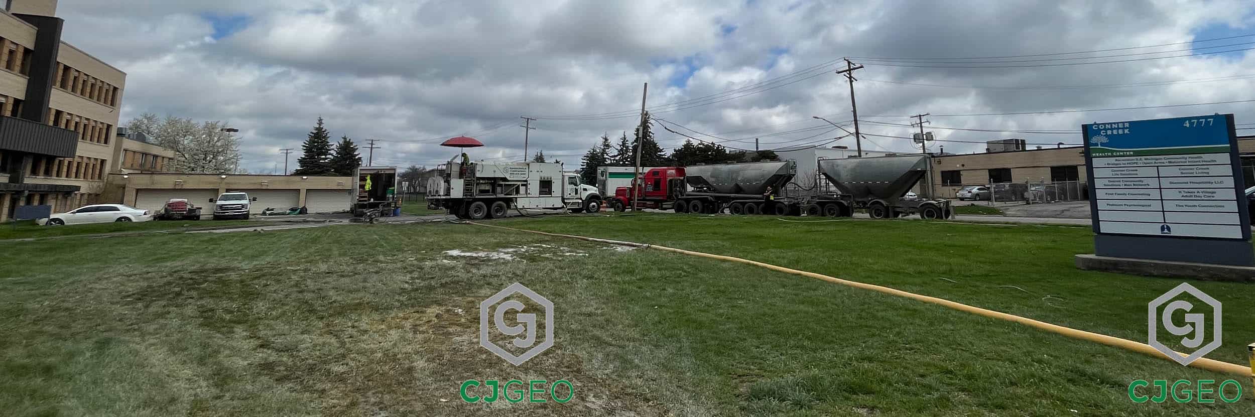 A CJGeo cellular grout plant, bulk cement tanker, and associated equipment sit adjacent to a manhole in a grassy field. The equipment is generating lightweight cellular concrete for annular grouting of a sewer slip line in Detroit, Michigan.