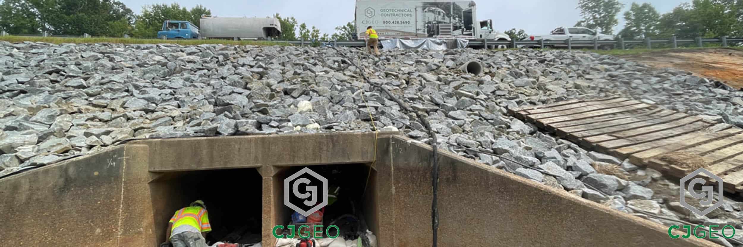 Two CJGeo crew members working on a box culvert repair. One crew member is inside the culvert, and the other is standing on the steep, riprap-covered embankment that extends up to the highway above.