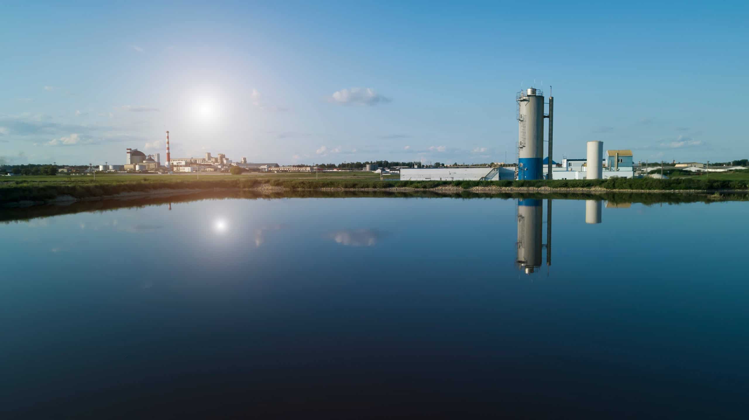 Industrial chemical or cement manufacturing facility reflected in a still retention pond under a clear blue sky — stationary emission source adjacent to surfac…Industrial chemical or cement manufacturing facility reflected in a still retention pond under a clear blue sky — stationary emission source adjacent to surface water requiring air quality permitting, stormwater compliance, and environmental impact assessment consulting by EDGE Engineering.