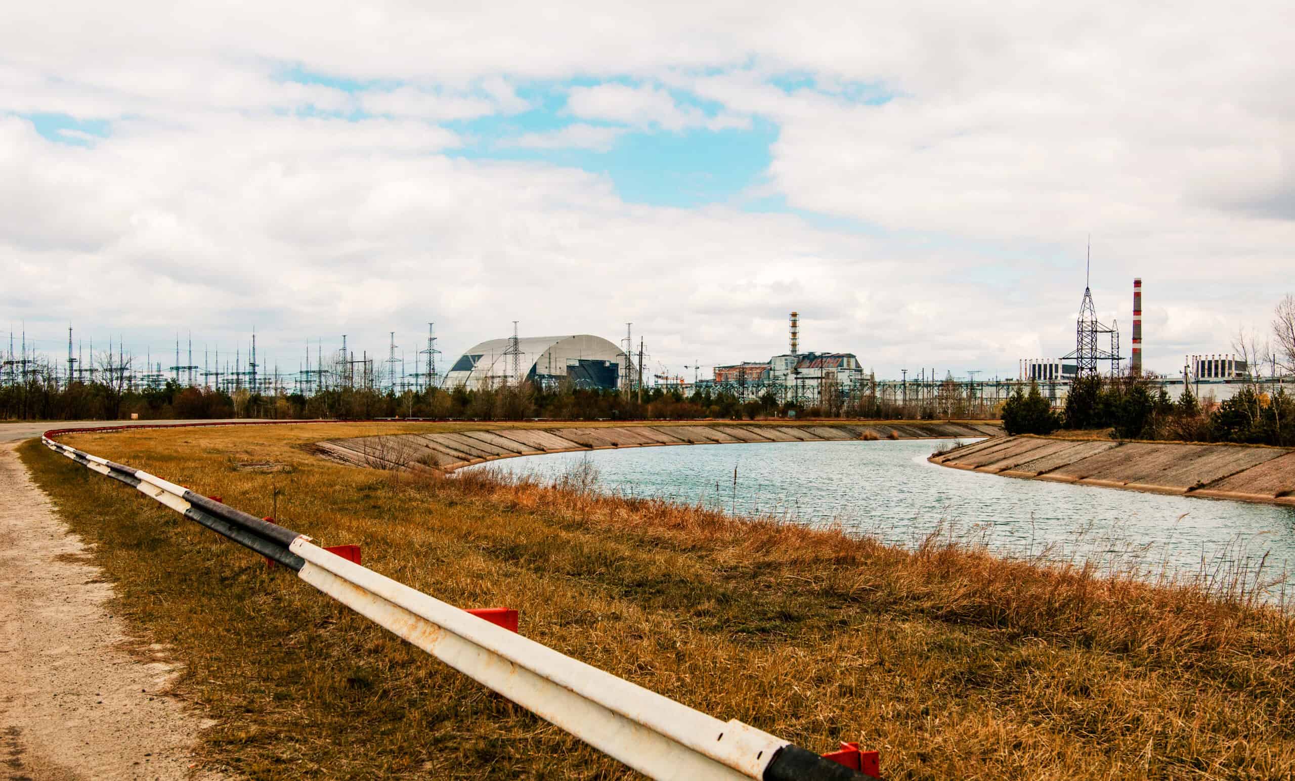 Chernobyl Nuclear Power Plant with its New Safe Confinement arch structure visible across an industrial canal, surrounded by flat terrain and power transmission infrastructure — site of the 1986 nuclear disaster used to illustrate industrial facility environmental risk and remediation challenges.