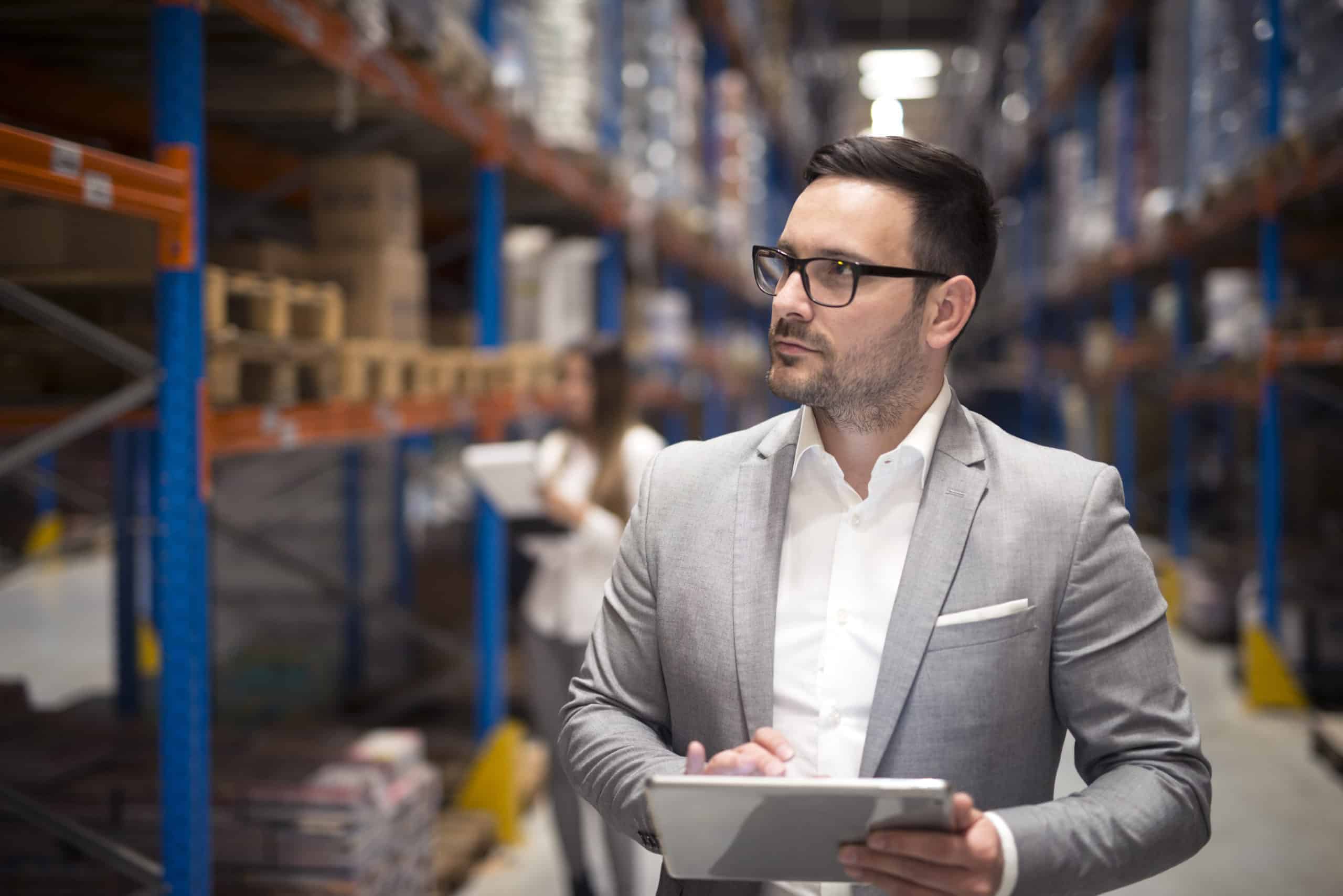 Portrait of successful businessman manager CEO holding tablet and walking through warehouse storage area looking towards shelves.