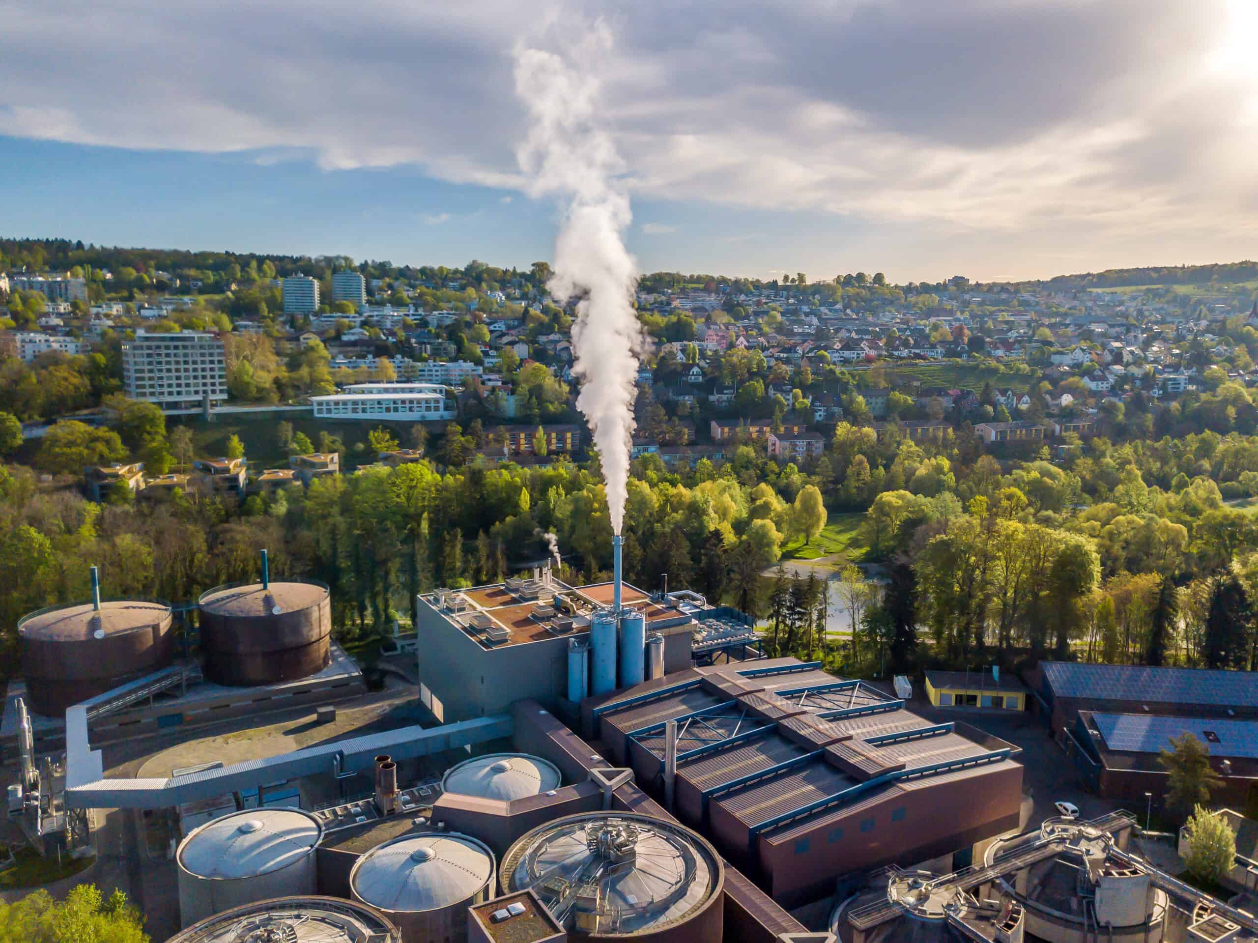 Aerial view of a wastewater treatment or industrial processing facility with digesters, storage tanks, and an active steam emissions stack adjacent to a reside…Aerial view of a wastewater treatment or industrial processing facility with digesters, storage tanks, and an active steam emissions stack adjacent to a residential community and green tree buffer — urban industrial facility subject to air quality permitting, odor control compliance, and environmental regulatory consulting by EDGE Engineering.