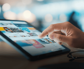 Uma mão toca a tela brilhante de um tablet, que exibe miniaturas de imagens coloridas, com óculos de leitura na mesa e luzes desfocadas ao fundo.