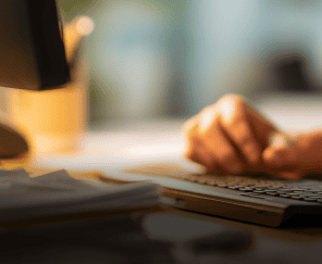 Uma pessoa trabalha em um computador, com as mãos sobre o teclado e documentos na mesa, em um ambiente de escritório com luz quente e focada.